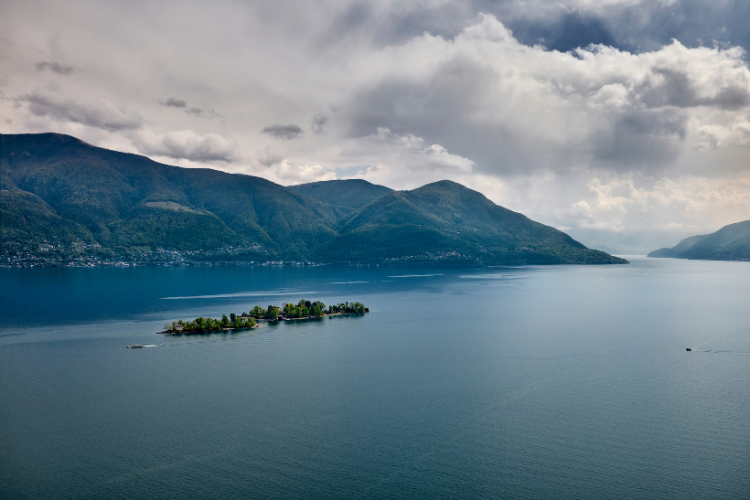 Kleine Insel im See vor einer weitläufigen Berglandschaft unter bewölktem Himmel.