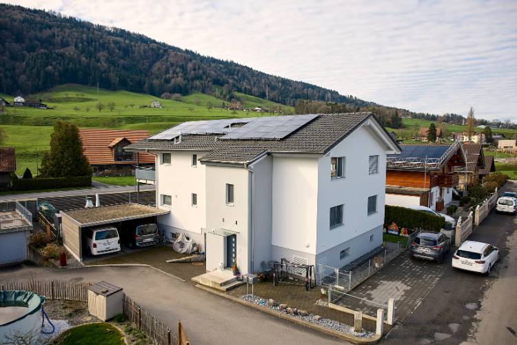 Ein modernes, weisses Wohnhaus mit Solarpanels auf dem Dach steht in einer ländlichen Umgebung neben einem Carport und vor grünen Hügeln.