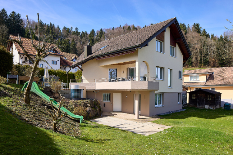 Modernisiertes Einfamilienhaus mit hellbeiger Fassade, braunem Satteldach, Balkon im ersten Stock, großem Garten und umliegender Wohngegend am Waldrand