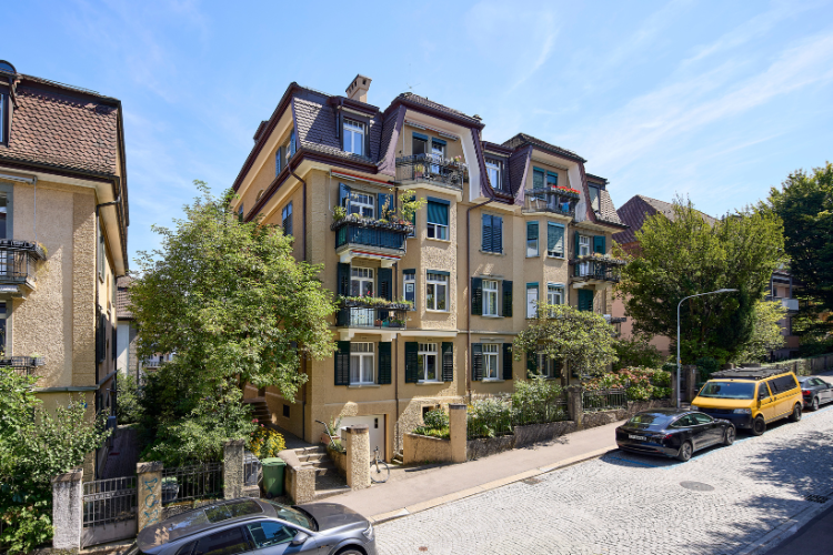 Edificio residenziale multipiano con facciata gialla, balconi e giardino verde in una tranquilla strada residenziale.