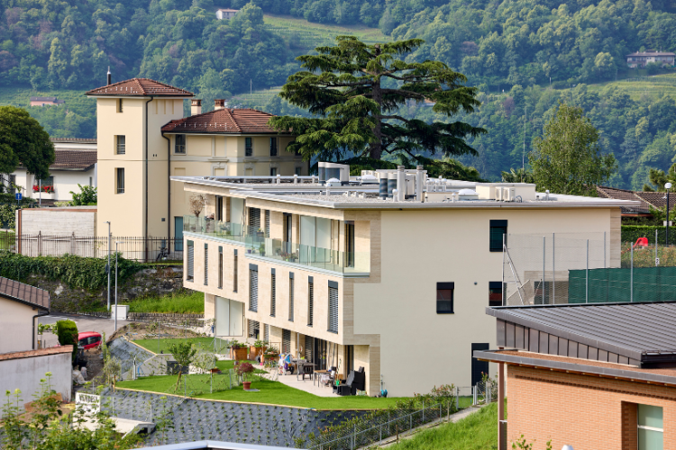 Moderno edificio plurifamiliare con ampi balconi in posizione collinare davanti a un paesaggio montano boscoso.