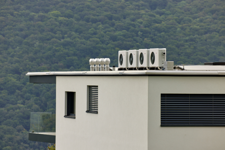 Ein modernes, würfelförmiges Wohnhaus mit grossen Fenstern und Balkonen steht auf einem grünen Hang vor einer bewaldeten Berglandschaft.