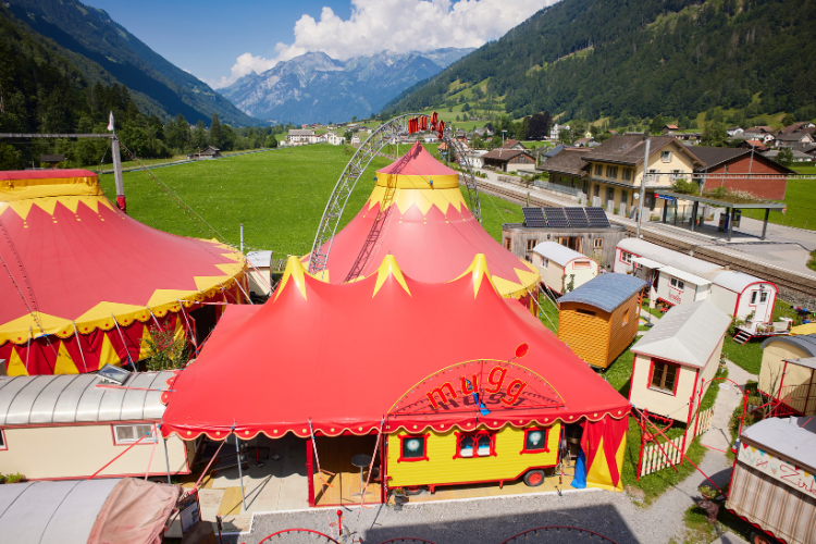 Buntes Zirkuszelt in Rot und Gelb mit umliegenden Wagen und grüner Berglandschaft im Hintergrund.