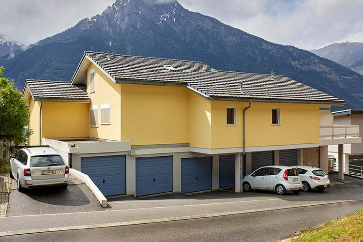 Ein gelb gestrichenes Wohnhaus mit mehreren blauen Garagentoren im Erdgeschoss, davor geparkten Autos und einer Berglandschaft mit hohen, teilweise schneebedeckten Gipfeln im Hintergrund.