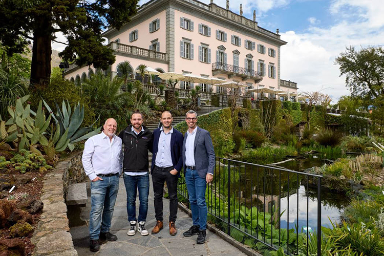 Vier Personen stehen auf einem Gartenweg vor einem grossen historischen Gebäude mit heller Fassade, umlaufenden Balkonen und einer üppigen Parkanlage mit Teich.
