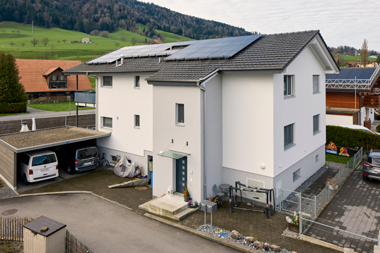 Ein modernes, weisses Wohnhaus mit Solarpanels auf dem Dach steht in einer ländlichen Umgebung neben einem Carport und vor grünen Hügeln.