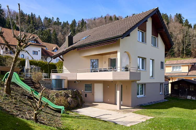Modernisiertes Einfamilienhaus mit hellbeiger Fassade, braunem Satteldach, Balkon im ersten Stock, großem Garten und umliegender Wohngegend am Waldrand