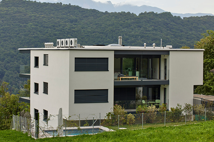 Ein modernes, würfelförmiges Wohnhaus mit grossen Fenstern und Balkonen steht auf einem grünen Hang vor einer bewaldeten Berglandschaft.