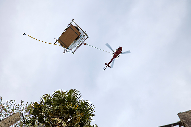 Ein roter Helikopter fliegt am Himmel und transportiert eine grosse, rechteckige Last, die an einem Seil unter dem Fluggerät hängt, während unten Palmen ins Bild ragen.