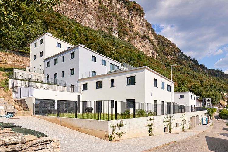 Mehrere moderne, weiss verputzte Wohngebäude stehen terrassenförmig an einem Hang unterhalb einer bewaldeten Felswand.