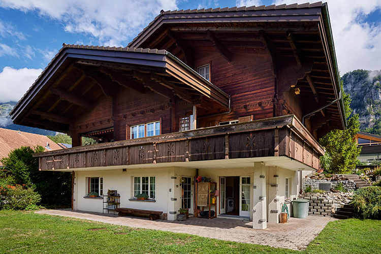 Ein traditionelles zweistöckiges Chalet aus Holz und Stein mit breitem Balkon und geschnitzten Details steht inmitten eines gepflegten Gartens vor einer bergigen Landschaft.