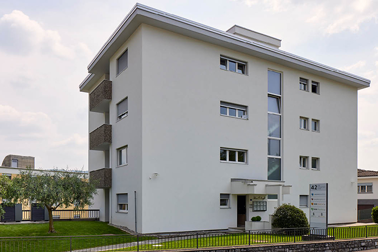 Ein modernes, helles Mehrfamilienhaus mit klaren Linien, mehreren Balkonen und einer schmalen vertikalen Glasfront steht auf einem gepflegten Grundstück mit Rasen und einem Baum im Vordergrund.
