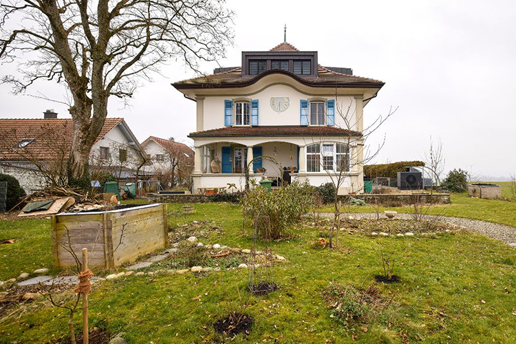 Ein zweistöckiges historisches Wohnhaus mit hellem Putz, blauen Fensterläden und einem markanten Dachaufsatz, umgeben von einem grossen Garten mit Beeten, jungen Bäumen und einem gewundenen Kiesweg. Neben dem Haus stehen weitere Wohngbäude, während im Hintergrunde offene Felder unter einem bewölkten Himmel zu sehen sind.
