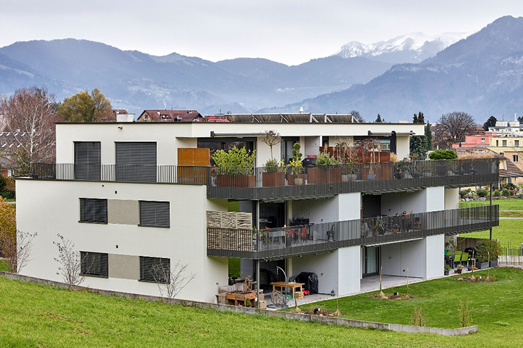 Ein modernes, flach gebautes Mehrfamilienhaus mit drei Etagen, hellen Fassaden und grossen umlaufenden Balkonen, die mit Pflanzen und Sitzbereichen gestaltet sind. Das Gebäude steht auf einer leicht abfallenden Wiese am Stadtrand, mit Blick auf angrenzende Wohngebäude, Bäume und eine Bergkulisse im Hintergrund.