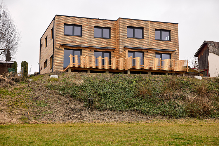 Ein modernes zweistöckiges Wohngebäude mit Holzfassade steht auf einer erhöhten Wiese und verfügt über eine breite Terrasse mit Holzgeländer.