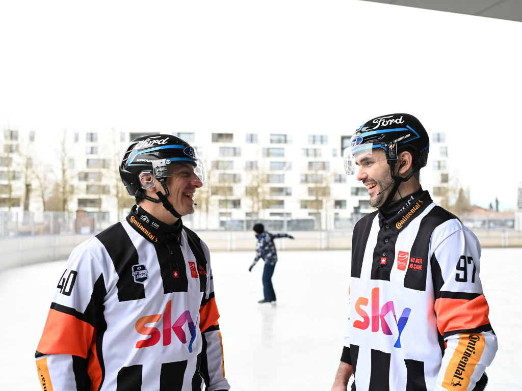 Zwei Eishockeyschiedsrichter des SHIF in Uniform lachen gemeinsam auf einer Eisbahn im Freien
