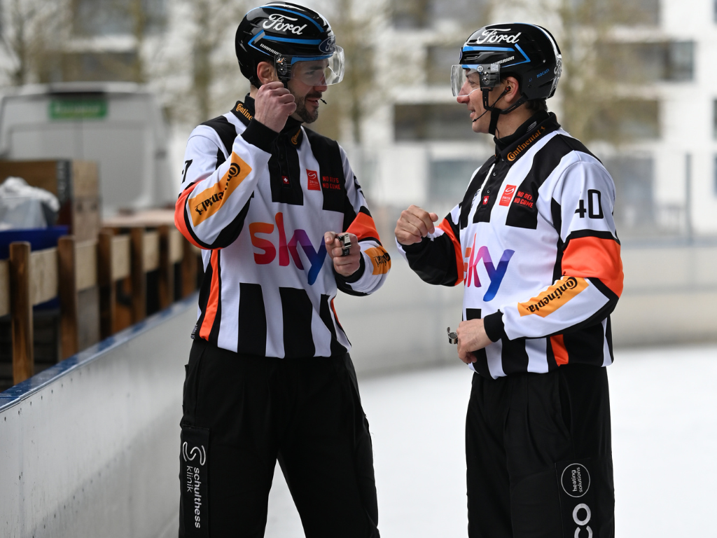 Zwei Eishockeyschiedsrichter des SHIF in Uniform besprechen ihr Vorgehen auf einer Eisbahn im Freien