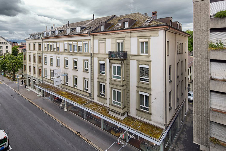 Mehrstöckiges Wohn- und Geschäftshaus mit heller Fassade an einer städtischen Strasse unter bewölktem Himmel.
