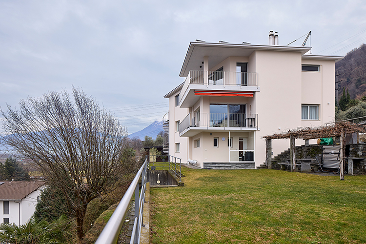 Modernes, helles Wohnhaus mit mehreren Balkonen und großem Garten, gelegen an einem Hang mit Bergblick.