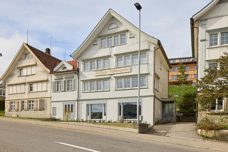 Historisches Gebäude mit heller Fassade und markanter Giebelfront an einer leicht ansteigenden Strasse.