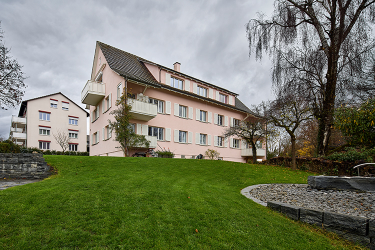Rosa Mehrfamilienhaus mit Balkonen und großem gepflegtem Garten unter bewölktem Himmel.