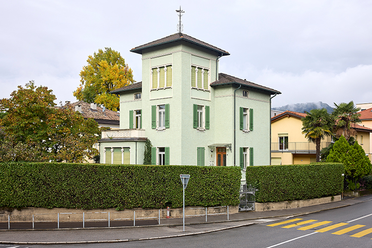 Hellgrünes Wohnhaus mit markantem Turm und grünen Fensterläden, umgeben von einer hohen Hecke an einer Strassenkreuzung.