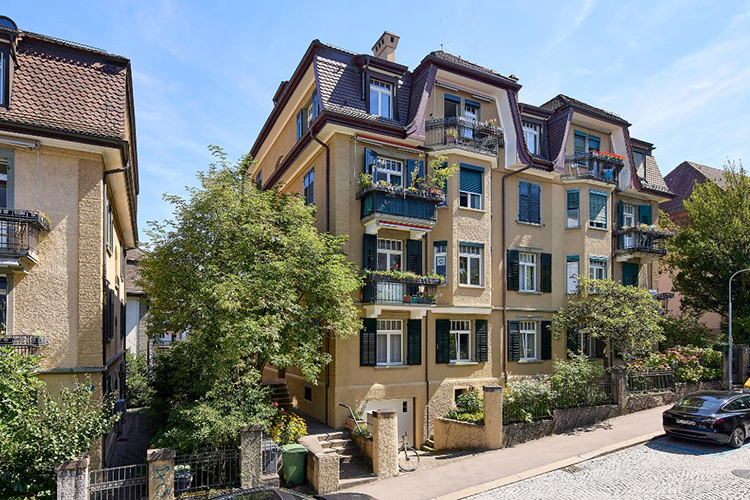 Mehrstöckiges Wohnhaus mit gelber Fassade, Balkonen und begrüntem Vorgarten in einer ruhigen Wohnstrasse.