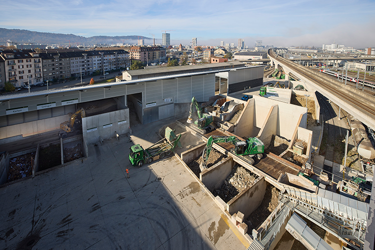 Recyclinganlage mit sortierten Materialboxen, Baufahrzeugen und angrenzender Bahntrasse in einem städtischen Industriegebiet.