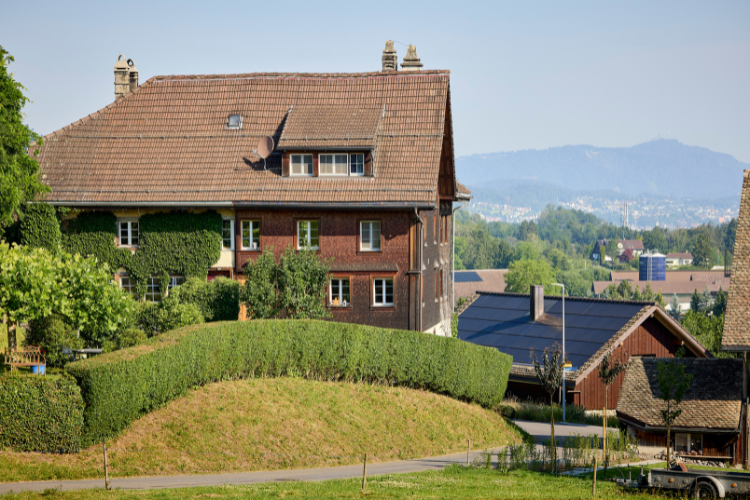 Traditionelles Wohnhaus mit Holzfassade und gepflegten Hecken vor einer hügeligen Landschaft.