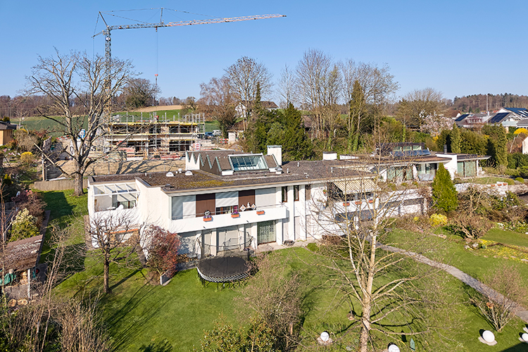 Modernes Flachdachhaus mit grossen Fenstern und weitläufigem Garten in leicht hügeliger Umgebung.