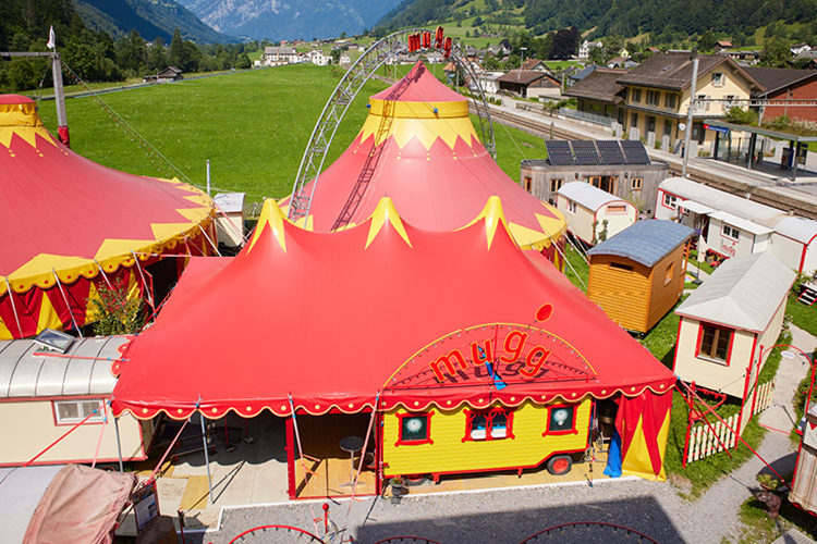Buntes Zirkuszelt in Rot und Gelb mit umliegenden Wagen und grüner Berglandschaft im Hintergrund.