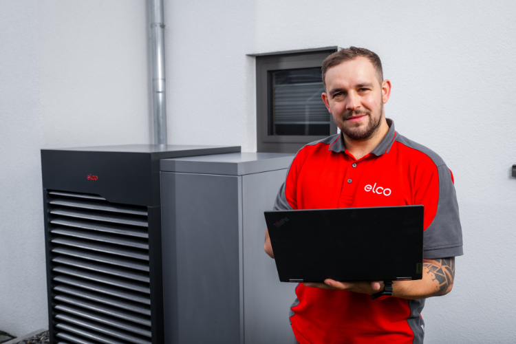 ELCO Servicetechniker in rotem Arbeitshemd steht vor einer modernen Wärmepumpe und blickt lächelnd in die Kamera.