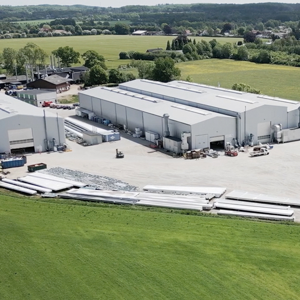 Vue aérienne de l'installation industrielle de Dencam, avec entrepôts, équipements et champs verdoyants environnants.