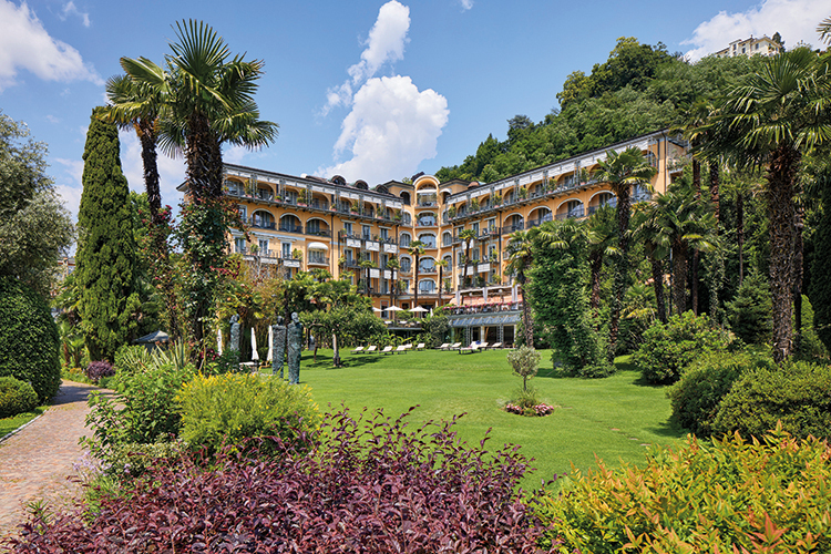 Ein grosses, elegantes Hotelgebäude mit gelber Fassade und vielen Balkonen, umgeben von üppigen Palmen, Büschen und einer weitläufigen grünen Gartenanlage, vor einem bewaldeten Hügel und blauem Himmel mit weißen Wolken.
